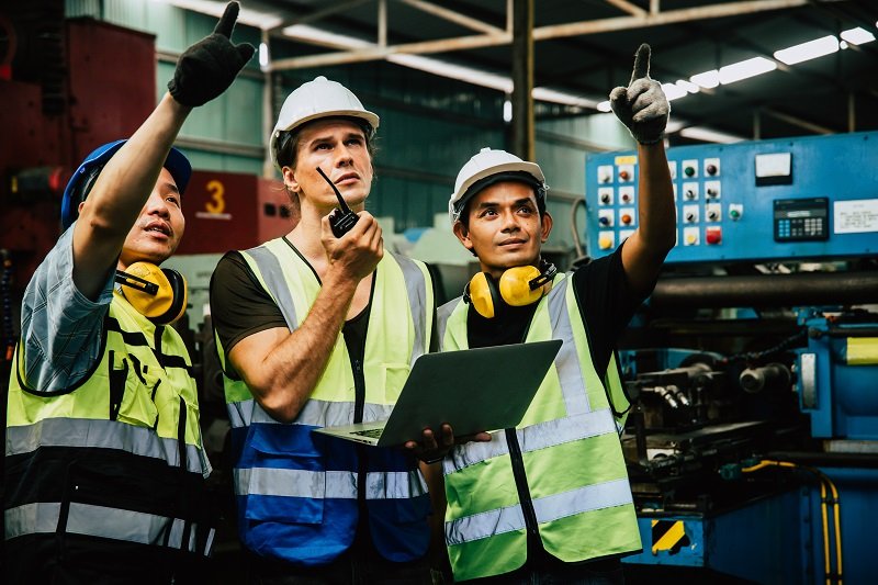 Três engenheiros no meio da indústria apontam para longe enquanto um deles fala ao rádio e segura um computador, representando a importância dos treinamentos para membros da CIPA