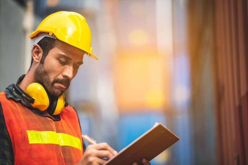 Homem usando capacete amarelo, colete e protetor auricular faz anotações, representando o que é avaliação de risco.