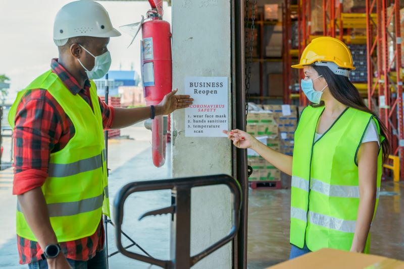 Dois trabalhadores em uma fábrica, usando capacetes, coletes sinalizadores e máscaras, apontam para um aviso sobre proteção contra coronavírus. 
