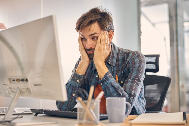 É permitido trabalhar 10h por dia? Homem cansado no escritório apoiando o rosto com as mãos.