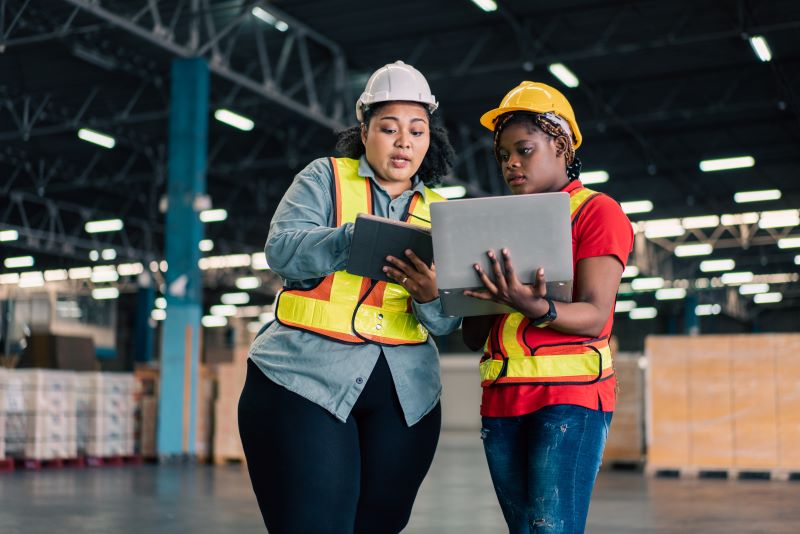 Engenharia de segurança do trabalho: duas mulheres analisando processos em armazém, vestindo EPIs e usando computador e tablet.